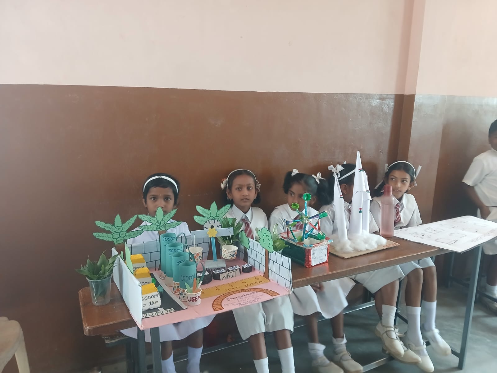 Schoolgirls sit beside their handmade environmental and measurement-themed projects on display.